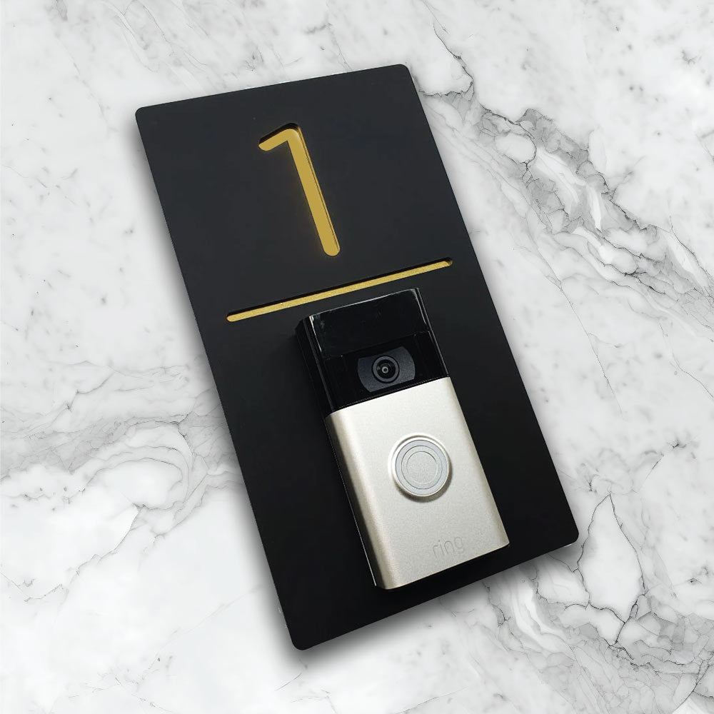Black vertical house sign with gold details with an added video ring doorbell on face of house plaque